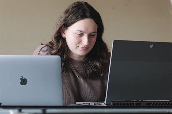 Photo of a student working at two computers, one with an Apple logo