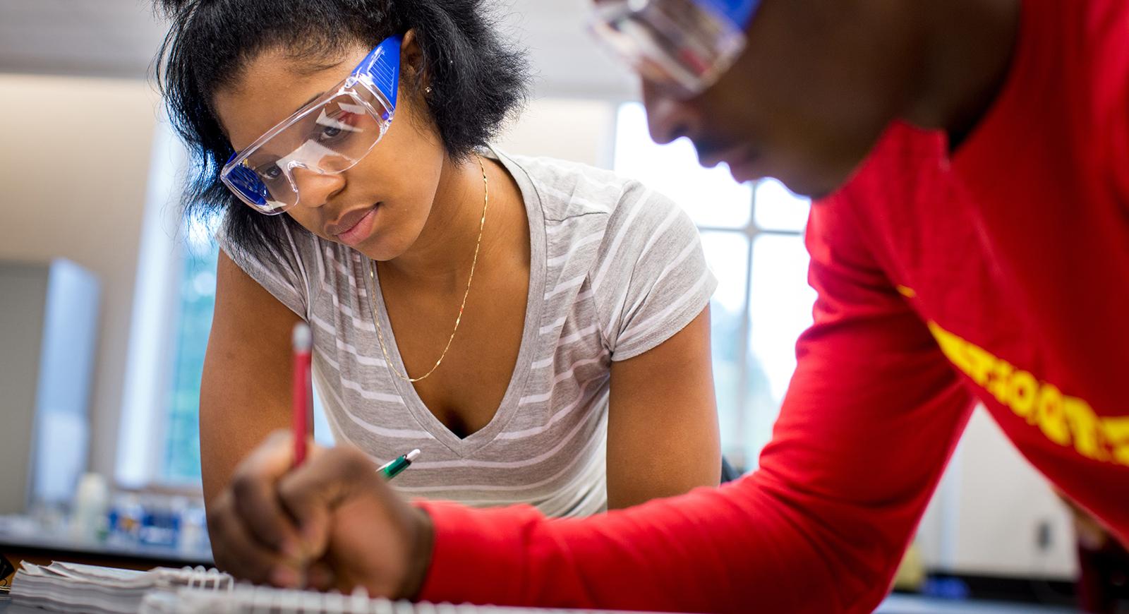 Photo of two Chatham University students working in a chemistry lab wearing protective goggles.