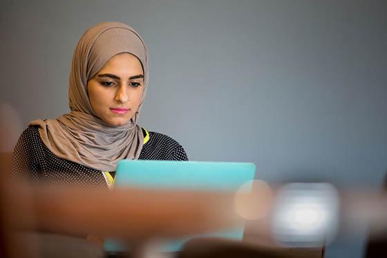 Photo of a Chatham University student wearing a hijab, smiling softly while working on her laptop in Cafe Rachel