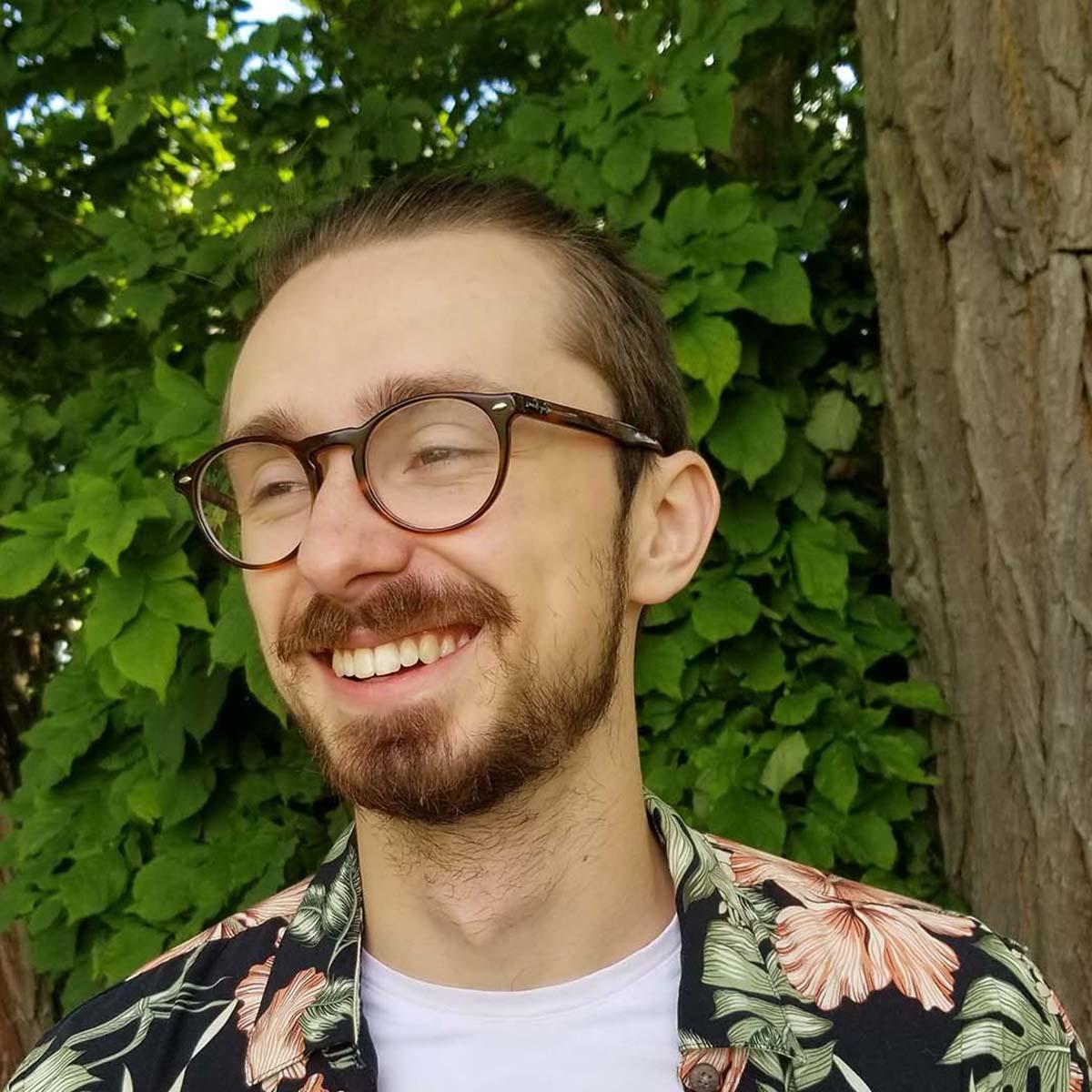 卡森·卡斯特的照片, 一个戴着眼镜，穿着鲜艳印花衬衫的白人年轻人, 在绿树前微笑