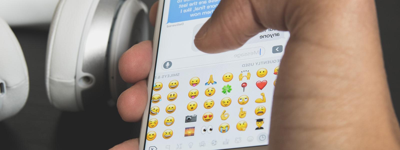 Close-up photo of a hand holding an iPhone while typing on an emoji keyboard.