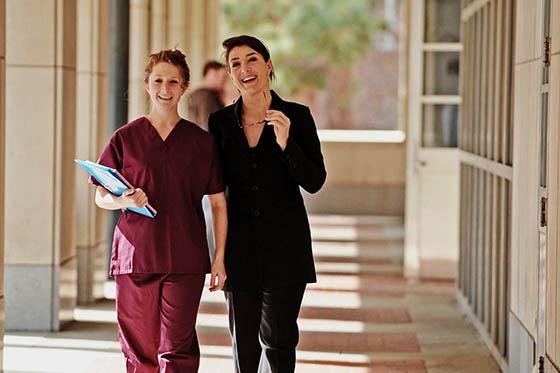 Photo of a nurse in maroon scrubs walking with a woman in a black suit in an outdoor hallway