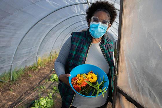 Photo of a Chatham University student in a mask, holding a bowl of harvested produce at the door of a greenhouse